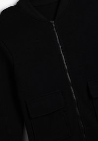Giacca nera a costine con zip frontale. Presenta un colletto rotondo e due grandi tasche frontali. Il materiale sembra avere una texture spessa.