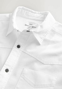 Hvid skjorte med struktureret stof, der har en brystlomme og sorte knapdetaljer. Etiketten siger "NEXT" med størrelse "M."