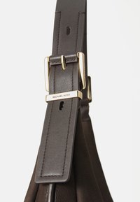 Bruine lederen band met een gladde textuur, voorzien van een gesp in goudkleur en een geperst logo. Het ontwerp bevat verstelgaten voor de maat.