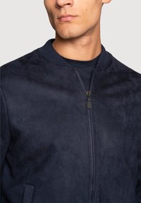 Chaqueta bomber navy hecha de suave ante con cuello acanalado, cierre frontal de cremallera y bolsillos laterales. Textura suave, diseño moderno.