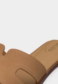 Lys brun skinn slide-sandal med utskjæringsdesign og synlige sømmer, på en nøytral bakgrunn.