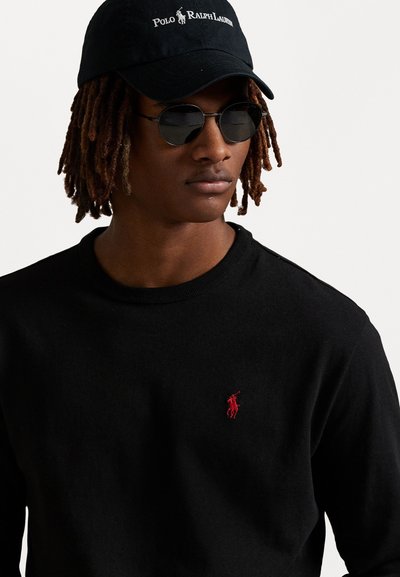 Sweatshirt de algodão preto com um decote redondo, apresentando um pequeno logótipo bordado em vermelho no peito. Combinado com óculos de sol pretos e um boné preto.