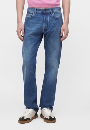 Personne portant un jean bleu droit, des baskets blanches et noires, et une chemise rose, debout devant un fond uni.