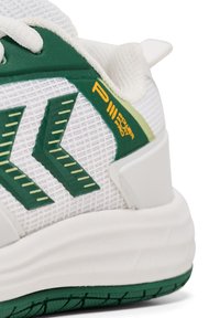 Vit sneaker med gröna accenter, texturerad mesh-överkropp och en dämpad sula. Anmärkningsvärda detaljer inkluderar ett geometriskt mönster och en logotyp på hälen.