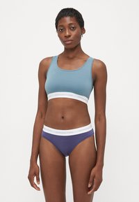 Nő kék sportmelltartóban és lila alsóneműben, fehér elasztikus pántokkal, melyekre az van írva, hogy "Copenhagen Studios", egy sima fehér háttér előtt.