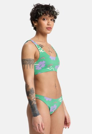 Frau mit kurzen lockigen Haaren in einem grünen zweiteiligen Badeanzug mit Blumendruck, die Arm- und Rumpftattoos zeigt, vor einem schlichten weißen Hintergrund stehend.