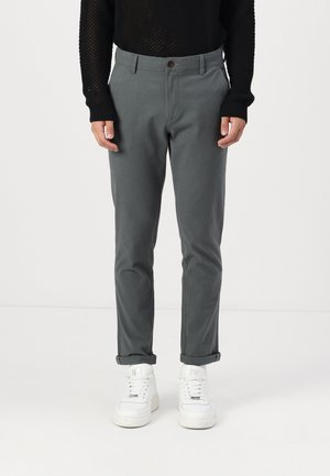 Pantalones de algodón grises ajustados, con cierre de botón y puños enrollados, combinados con zapatillas blancas. Suéter de punto texturizado arriba.