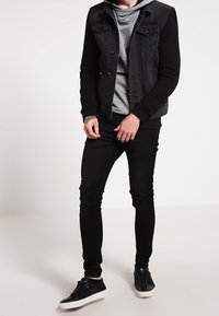 Man draagt een zwarte spijkerjas, een grijze hoodie, zwarte skinny jeans en zwarte sneakers met witte zolen, staand tegen een witte achtergrond.