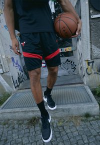 Sorte basketballshorts med røde striber og logo, parret med sorte Nike-sko med hvide detaljer, mens en basketball holdes i den ene hånd.