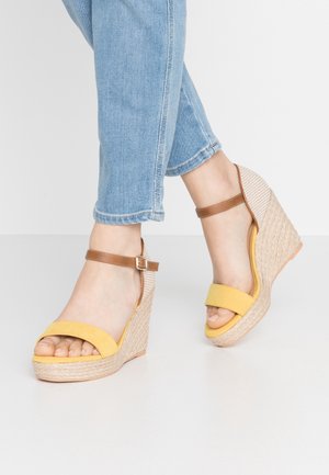 Fødder i blå jeans iført gule og tan kile-sandaler med ankelremme og flettede platformhæle på hvidt gulv.
