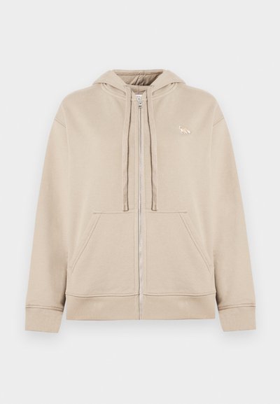 Felpa beige con zip realizzata in tessuto morbido, dotata di tasca a marsupio, cappuccio con coulisse e un piccolo logo accentato sul petto.