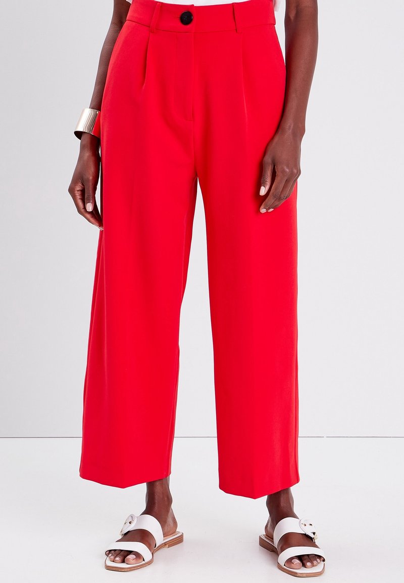Pantalon rouge à jambes larges en tissu lisse ; doté de plis avant, d'un seul bouton noir et d'une taille ajustée. Porté avec des sandales blanches.