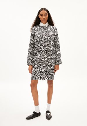 Schwarz-weiß gemustertes Kleid mit langen Ärmeln, Kragen und einem leicht lockeren Schnitt. Kombiniert mit weißen Socken und schwarzen Schuhen.