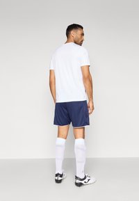 Balti sportinė marškinėliai, mėlyni šortai su baltais kraštais, balti per kelio aukščio kojinės ir juodi futbolo bateliai. Vaizdas iš nugaros prieš šviesią fono.