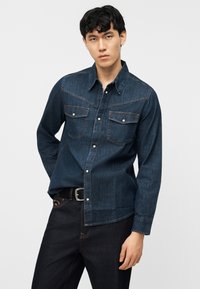 Νεαρός άνδρας που φοράει ένα μπλε σκούρο τζιν πουκάμισο με κουμπιά και μπροστινές τσέπες, καθώς και μαύρο τζιν παντελόνι, στέκεται μπροστά σε ένα απλό ανοιχτό φόντο.