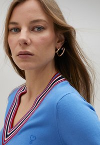 Blå V-ringad tröja med flerfärgad randning i röd, vit och marinblå. Hjärtbroderi på bröstet. Texturerad stickad tyg. Guldörhängen i form av hoops.