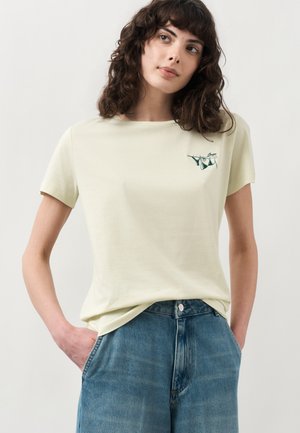 Kvinde med krøllet mørkt hår iført en blege gul T-shirt med en lille undertøjslinjegrafik og blå jeans, hænderne i lommerne.