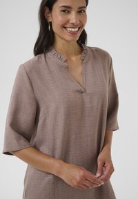 Svijetlosmeđa bluza s opuštenim krojem, polu rukavima i nabranim izrezom. Tkanina ima suptilnu teksturu. Model se smije.