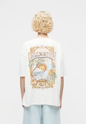 Osoba s kučeravými blond vlasmi nosiaca bielu oversized tričko s farebným tropickým motívom loga Billabong a nápisom "1973" na zadnej strane.