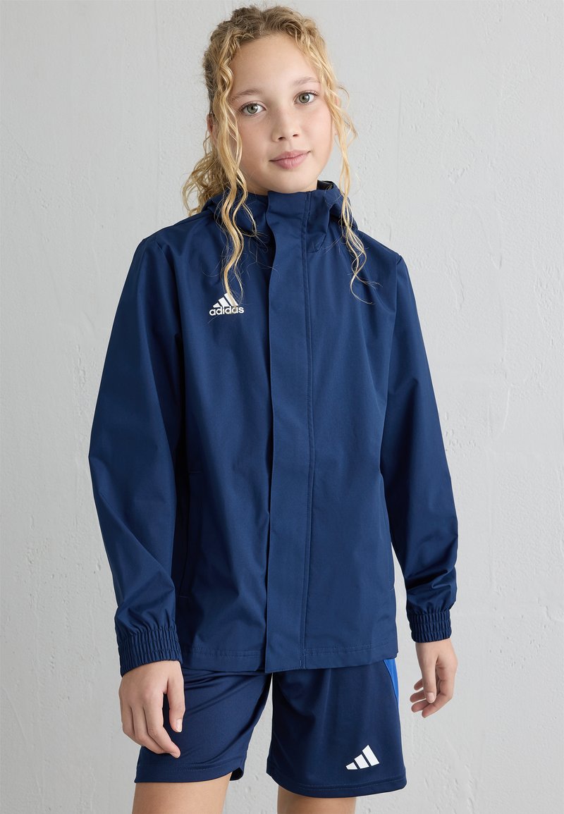 Tums zils ūdensizturīgs jaciņa ar augstu apkakli un elastīgām piedurknēm, ar baltu Adidas logo. Saskaņota ar atbilstošām tumši ziliem šortiem.