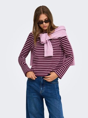 Femme portant des lunettes de soleil noires, une chemise à manches longues rayée violette et blanche, un jean bleu et un pull violet clair posé sur ses épaules.