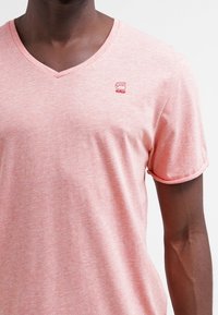 Jasnoróżowa, krótko rękawowa bawełniana koszulka z dekoltem w serek oraz małym czerwonym logo na piersi. Miękka tekstura z melanżowym wzorem.