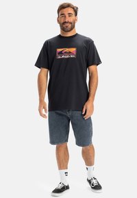 Sort t-shirt med et farverigt Quiksilver-logo, parret med denim shorts og sorte sko. Har korte ærmer og en afslappet pasform.