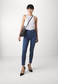 Un jean en denim bleu taille haute associé à un débardeur blanc et des chaussures à talons noires. Un petit sac noir est porté à l'épaule.