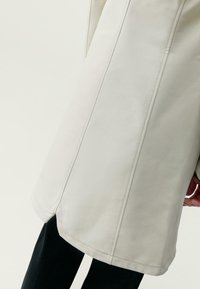 Svetlo béžový kabát vyrobený z hladkej tkaniny, s vertikálnymi švami, priamym strihom a postrannými rozparkami na spodnom okraji pre voľnosť pohybu.