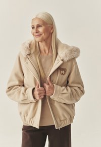 Beżowa kurtka z kapturem obszytym sztucznym futerkiem, zapinana na zamek, z naszywką w kształcie misia. Miękka w dotyku, o luźnym kroju, z ściągaczami na mankietach i u dołu.