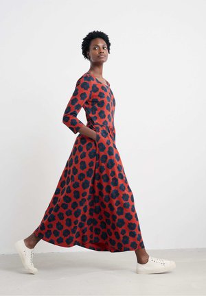 Kvinde går i en rød maxi-kjole med sorte blomster mønstre og hvide sneakers mod en ensfarvet hvid baggrund.