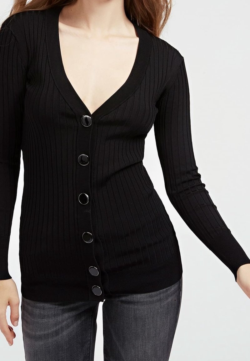 Femme portant un cardigan noir ajusté, côtelé, boutonné, à manches longues, et un jean gris foncé, montré du cou aux hanches.