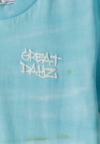Koton majica v svetlo modri barvi z vzorcem tie-dye. Vsebuje vezeno logo "GREAT DAYZ" v beli barvi. Teksturovana tkanina z rebrastim ovratnikom.