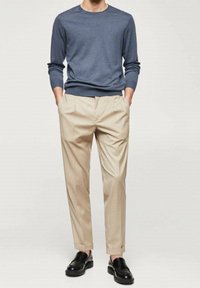 Mann in einem blauen Langarmhemd, beigen, gefalteten Hosen mit Umschlägen, schwarzen Lederloafers, der mit den Händen in den Taschen steht.