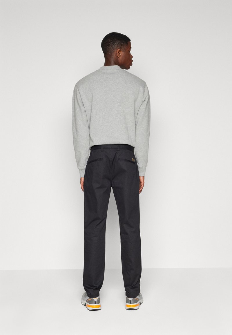 Grauer Sweatshirt mit langen Ärmeln, kombiniert mit dunklen Hosen. Zu den hervorstechenden Merkmalen gehören ein Rundhalsausschnitt und ein Logo-Patch auf den hinteren Taschen.