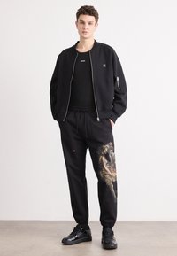 Black bomber jacket with a zip closure over a black t-shirt; black sweatpants featuring a colorful artwork detail and elastic cuffs; black sneakers.