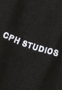 Zwarte stof met een gladde textuur, waarop de tekst "CPH STUDIOS" in witte geborduurde letters staat, gecentreerd en in vetgedrukte lettertype.