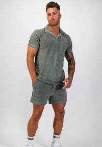 Khakigrön, kortärmad polo och matchande shorts i mjukt frottétyg; har en ficka på vänster bröst. Vita randiga strumpor och sneakers.