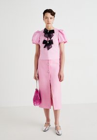 Kvinne iført en rosa puffermet topp med svarte sløyfer, rosa knappet midiskjørt med splitt, som holder en rosa frynseveske og sølvfargede sko.