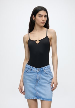 Frau mit langen dunklen Haaren, die ein schwarzes ärmelloses Top mit dünnen Trägern und einen hellblauen Jeansrock vor einem schlichten Hintergrund trägt.