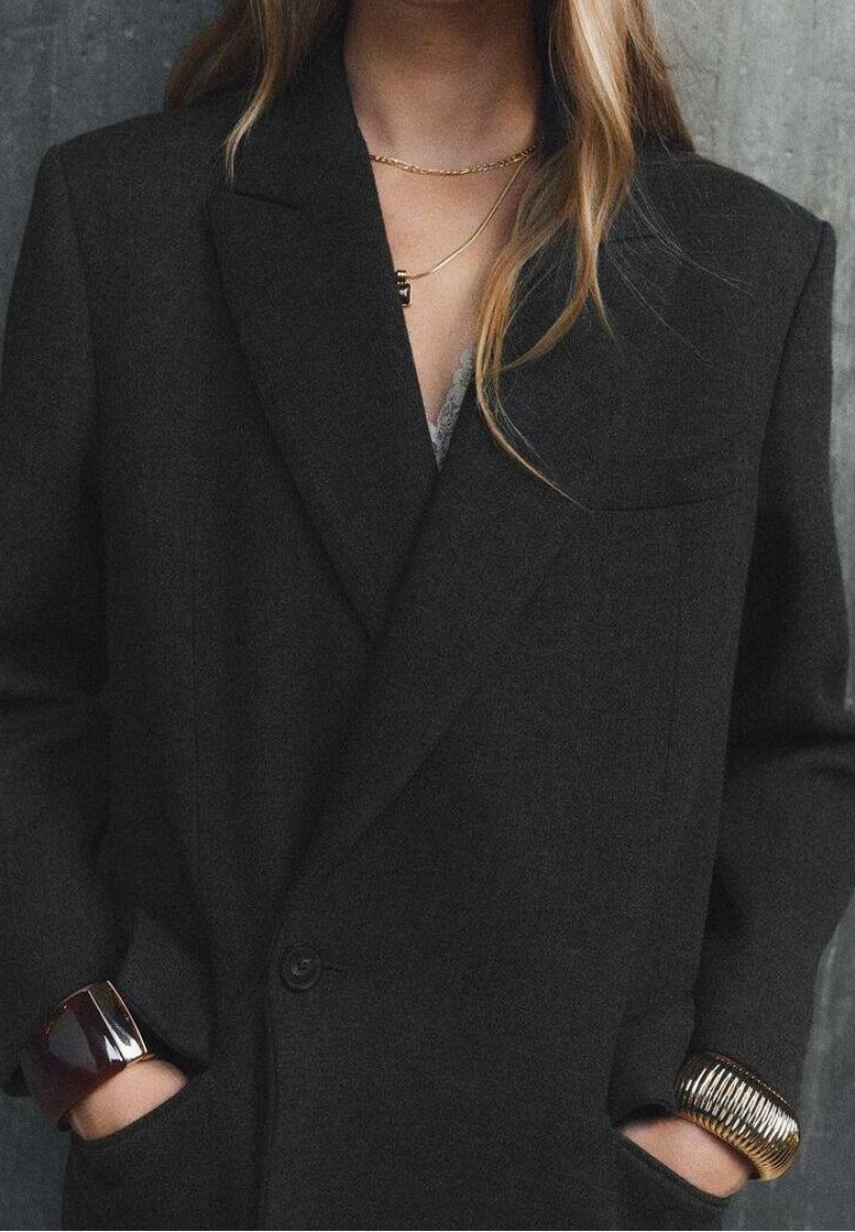 Femme portant un blazer sombre oversize, bracelets dorés et foncés, colliers dorés, mains dans les poches du blazer sur un fond gris texturé.