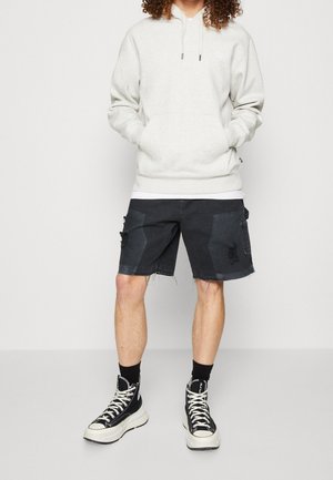 Persoon draagt een lichtgrijze hoodie, zwarte denim shorts met patches, zwarte sokken en zwart-witte hoge sneakers, staand met handen in de zakken.