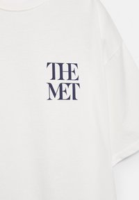 T-shirt blanc à manches courtes avec texte en serif bleu marine « THE MET » imprimé sur la partie supérieure gauche de la poitrine.