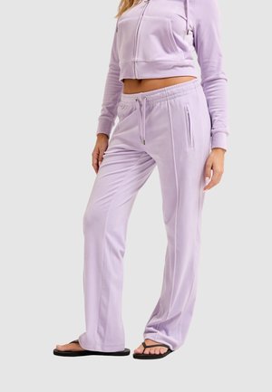 Vrouw met lila trainingsbroek en bijpassende korte zip-up hoodie met zwarte slippers, staand tegen een effen achtergrond.