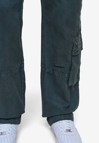 Spodnie cargo w ciemnozielonym kolorze z wieloma kieszeniami, widocznym przeszyciem i nieco luźniejszym krojem. W zestawie z białymi sneakersami.