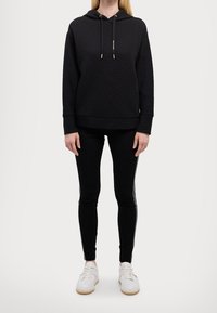 Felpa nera trapuntata con coulisse e tasca frontale, abbinata a leggings neri con accenti laterali e sneakers bianche.