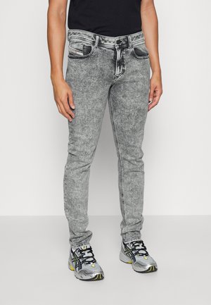 Personne portant un jean gris slim fit délavé à l'acide avec poches avant, bouton noir et chaussures de sport grises avec des détails noirs et jaunes.