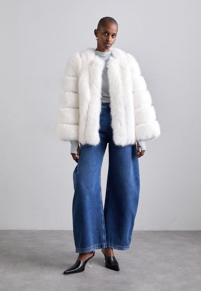 Weiße Kunstfell-Cropped-Jacke mit horizontalen Streifen, kombiniert mit hoch taillierten, weit geschnittenen blauen Jeanshosen und schwarzen spitzen Absätzen.
