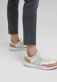 Zapatillas de deporte blancas, beige y rosas con cordones, combinadas con unos jeans oscuros recortados, sobre un fondo gris neutro.