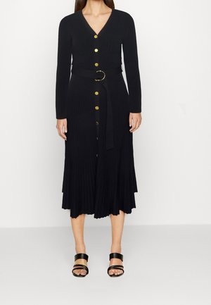 Kobieta w czarnej, długiej sukience midi z guzikami i dopasowanym paskiem, nosząca czarne, otwarte sandały na obcasie.